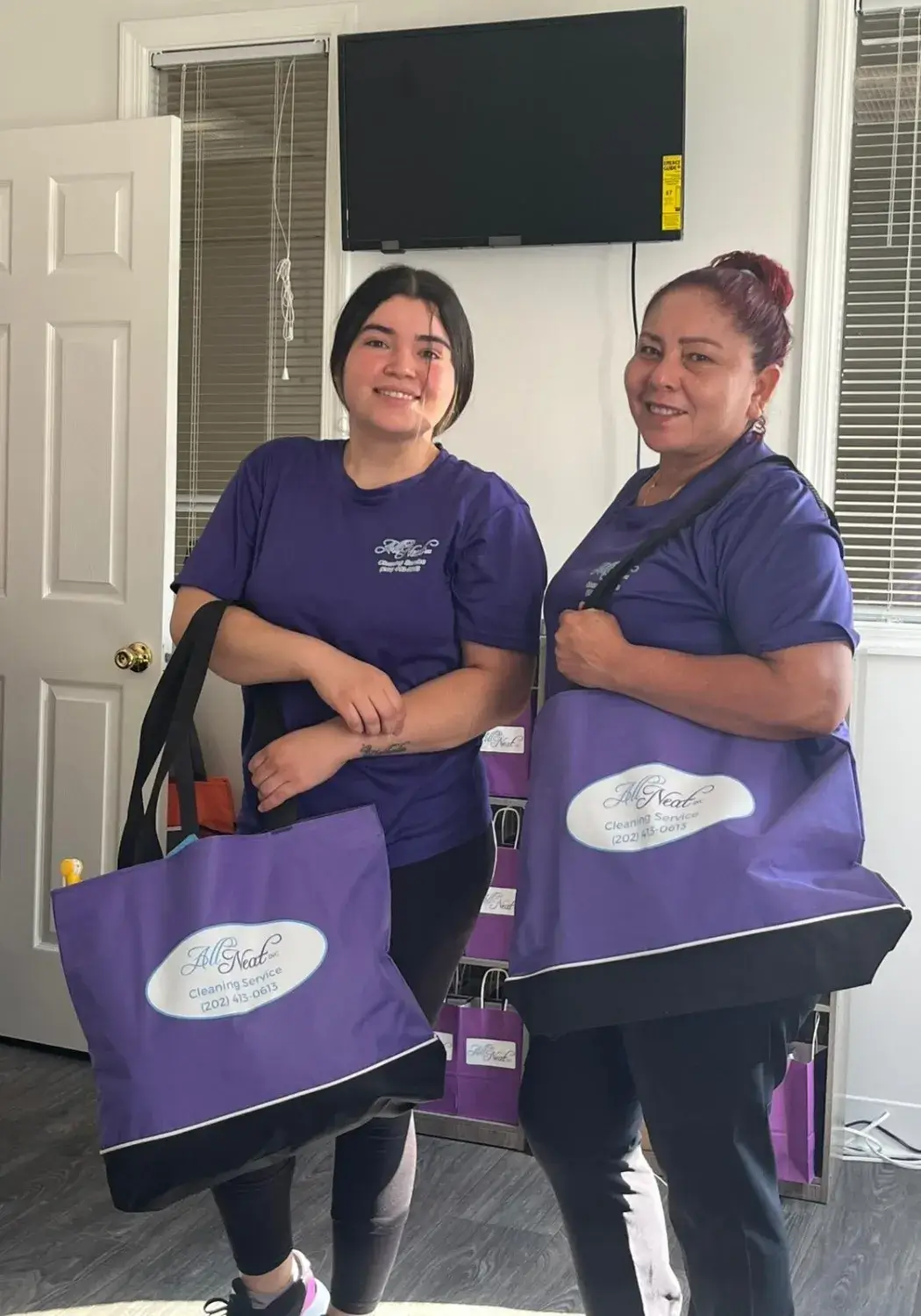 Two All Neat Cleaning Services employees smiling and holding branded purple cleaning supply bags